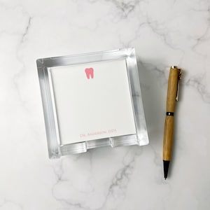 May include: A clear acrylic notepad holder with a white notepad featuring a pink tooth graphic and the text "DR. ANDERSON, DDS." A wooden pen with gold accents rests beside the notepad.