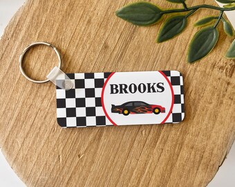 May include: A rectangular keychain featuring a black and white checkered pattern on the left and a white circle with a red border on the right. Inside the circle is the name BROOKS above a black race car with orange flame graphics.