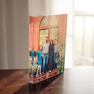 May include: A family of four poses for a photo in front of a brick building with a large arched window. The family is wearing blue clothing and smiling. The photo is printed on a clear acrylic block.