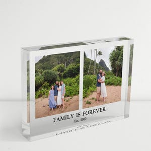 May include: A clear acrylic photo block displaying two family photos. The block features the text "FAMILY IS FOREVER Est. 2010" at the bottom. The photos show families in a tropical setting.