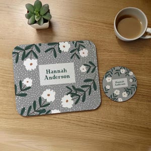 May include: A gray and white floral design coaster set with the name "Hannah Anderson" printed on both the coaster and the larger mat. The coaster set features white flowers, green leaves, and a gray background with white dots.