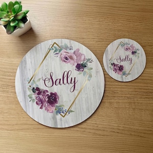 May include: A set of two round wooden coasters with a white woodgrain background. The coasters feature a gold geometric frame with purple and pink floral designs. The name "Sally" is written in cursive inside the frame on both coasters.