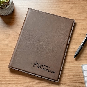 May include: A brown rectangular notebook with a textured cover featuring the name Jessica Anderson engraved in black script and block lettering. The notebook is positioned on a flat surface next to a black retractable ballpoint pen.