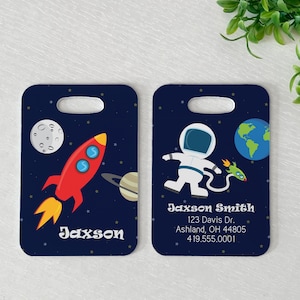 May include: Two blue luggage tags with a space theme. One tag features a red rocket ship with a blue and white moon in the background. The other tag features a white astronaut with a green rocket ship and a blue and green planet in the background. Both tags have the name "Jaxson" printed on them.