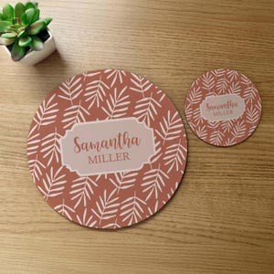 May include: A set of two round placemats with a terracotta background and a white leaf pattern. The larger placemat has a white rectangle with the text "Samantha MILLER" in a cursive font. The smaller placemat has the same pattern and the text "Samantha MILLER" in a smaller white rectangle.
