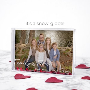 May include: A clear acrylic snow globe with a family photo inside. The photo shows a family of five sitting on a tree root by a lake. The text "Family is Forever" is printed on the base of the snow globe, along with the names "Mark, Susan, Olivia, Evie & James".