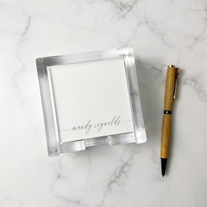 May include: A square clear acrylic memo holder containing a stack of white paper sheets with the name mandy reynolds printed in a dark script font, accompanied by a light brown wooden ballpoint pen with black metal accents.