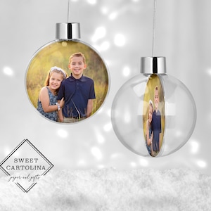 May include: Two clear glass Christmas ornaments with silver tops. Each ornament has a photo of a young boy and girl smiling at the camera. The photo is printed on a round piece of paper inside the ornament.