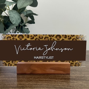 May include: A brown acrylic desk sign with a gold glitter leopard print design. The sign reads "Victoria Johnson, Hairstylist".