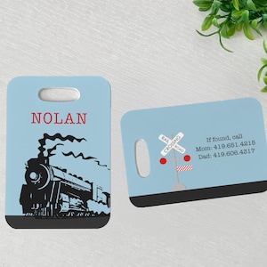 May include: Blue luggage tag with a black and white illustration of a train with smoke. The tag has a name tag that says "NOLAN". The back of the tag has a blue background with a red and white railroad crossing sign and the text "If found, call Mom: 419.651.4215 Dad: 419.606.4317"