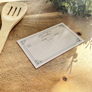 May include: A white recipe card with a black border and decorative flourishes. The card has lines for writing down a recipe, including the recipe name, servings, prep time, cook time, and ingredients. The text "Made with love by Grandma" is printed at the top of the card.