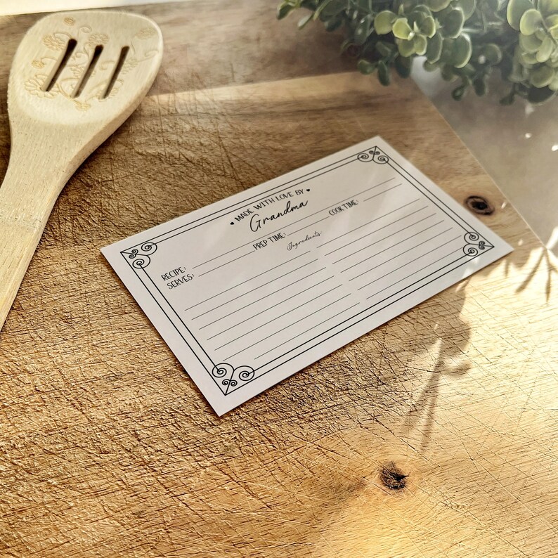 May include: A white recipe card with a black border and decorative flourishes. The card has lines for writing down a recipe, including the recipe name, servings, prep time, cook time, and ingredients. The text "Made with love by Grandma" is printed at the top of the card.
