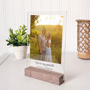 May include: A clear acrylic photo plaque set in a rectangular wooden base. The plaque features a printed photograph of a couple standing in a field at sunset, with the text "You're my favorite" and "CHAD & JESSICA" printed in black below the image.