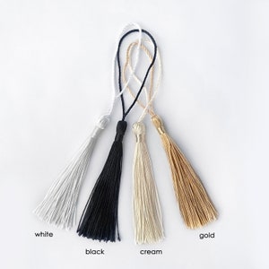 May include: Four decorative tassels in white, black, cream, and gold. Each tassel has a looped cord for hanging. The colors are labeled with the corresponding color name at the bottom of the image.