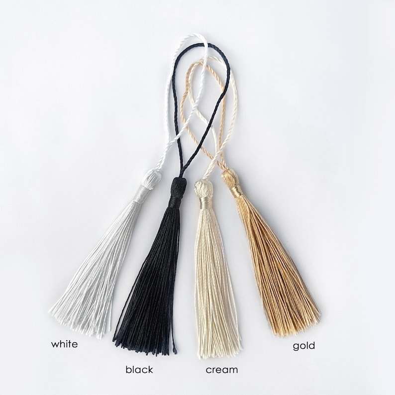 May include: Four decorative tassels in white, black, cream, and gold. Each tassel has a looped cord for hanging. The colors are labeled with the corresponding color name at the bottom of the image.