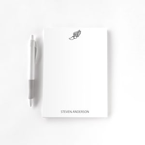 May include: A white notepad with a black winged shoe logo and the name "STEVEN ANDERSON" printed at the bottom. A silver and white pen rests on the left side of the notepad.