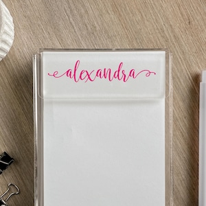 May include: A clear acrylic notepad holder with a white pad and a pink cursive "alexandra" on the top. A white pen and two black binder clips are also visible. The notepad is on a light brown wooden surface.