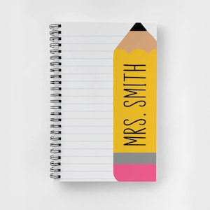 May include: A yellow pencil with a pink eraser and the text "MRS. SMITH" written on it. The pencil is on a white notebook with blue lines.