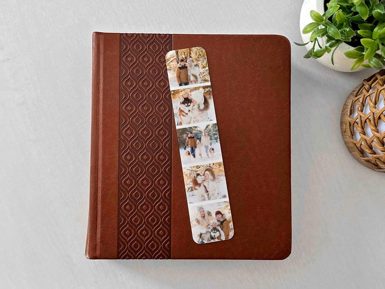 May include: A brown leather-bound journal with a patterned spine, and a photo bookmark. The bookmark displays a series of family photos. A small potted plant and woven basket are in the background.