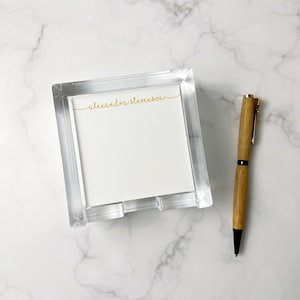 May include: A clear acrylic notepad holder with a white notepad and a wooden pen. The notepad has the name "alexandra stevenson" in gold script. The pen has a gold clip and black ink.