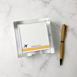 May include: Clear acrylic notepad holder with a white notepad featuring a blue airplane graphic and the name "ANDREW SMITH" above yellow stripes. A wooden pen with gold accents rests beside the notepad.