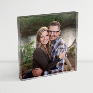 May include: A couple poses for a photo in a wooded area. The woman is wearing a brown sweater and the man is wearing a plaid shirt. They are both smiling and looking at the camera.