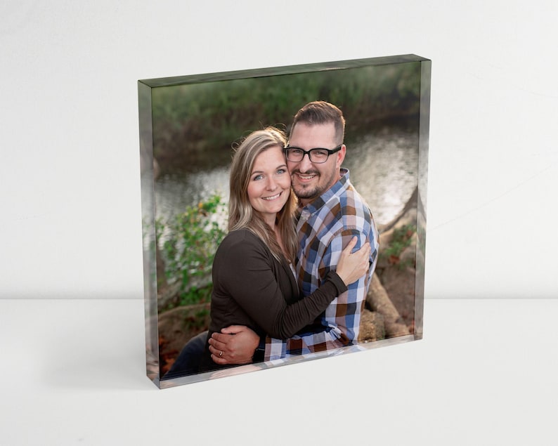 May include: A couple poses for a photo in a wooded area. The woman is wearing a brown sweater and the man is wearing a plaid shirt. They are both smiling and looking at the camera.