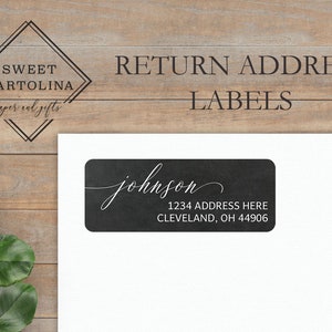 May include: A black return address label with the name "johnson" in elegant script. The label also includes the address "1234 ADDRESS HERE, CLEVELAND, OH 44906". The label is on a white background, with a wooden surface in the background.