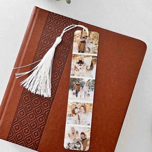 May include: A personalized bookmark featuring a series of family photos, with a white tassel attached. The bookmark is displayed on a brown leather-bound book with a decorative pattern. The photos show people with dogs in a snowy environment.