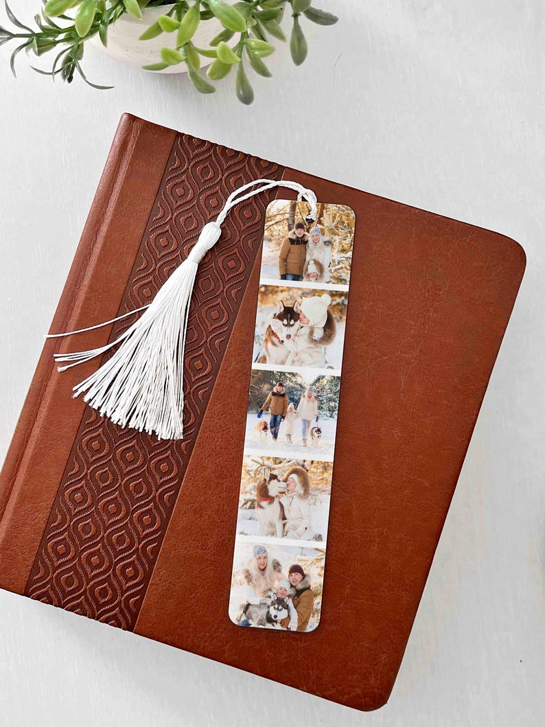 May include: A personalized bookmark featuring a series of family photos, with a white tassel attached. The bookmark is displayed on a brown leather-bound book with a decorative pattern. The photos show people with dogs in a snowy environment.