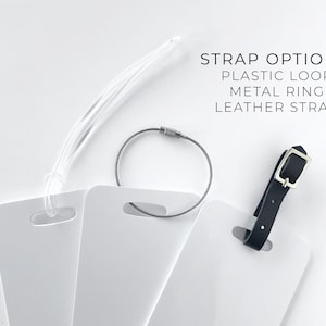 May include: White luggage tags with three strap options: a clear plastic loop, a gray metal ring, and a black leather strap. The text "STRAP OPTIONS: PLASTIC LOOP METAL RING LEATHER STRAP" is displayed.