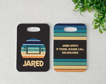 May include: Two rectangular luggage tags with rounded corners and a top cutout. The left tag features a gaming console graphic over a striped sunset circle and the name JARED. The right tag displays striped patterns and the text JARED SMITH IF FOUND, PLEASE CALL 555.555.5555.
