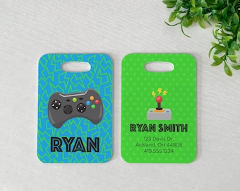 May include: Two rectangular luggage tags. One is blue with a green lightning bolt pattern and a black game controller. The other is green with a joystick graphic and the name "RYAN SMITH" and address.