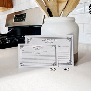 May include: Two blank recipe cards with a black and white decorative border. The larger card is 4x6 inches and the smaller card is 3x5 inches. Both cards have the text "Made with Love By" and a space to write a name. The cards are on a white countertop.