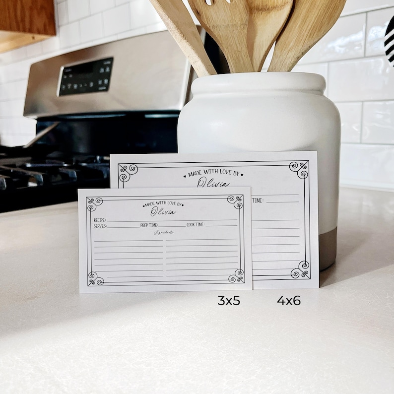 May include: Two blank recipe cards with a black and white decorative border. The larger card is 4x6 inches and the smaller card is 3x5 inches. Both cards have the text "Made with Love By" and a space to write a name. The cards are on a white countertop.