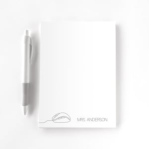 May include: A white notepad with a minimalist black line drawing of a computer mouse and the text "MRS ANDERSON". A silver and gray pen rests beside the notepad. The design is simple and modern.