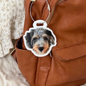 May include: A white luggage tag with a photo of a dog's face, attached to a brown backpack with a silver wire loop. The dog has black, gray, and tan fur. The backpack has a zipper and a front pocket.