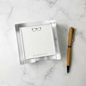May include: A clear acrylic notepad holder with a white notepad featuring a black eyeglasses graphic and the text "DR. ANDERSON, O.D." A wooden pen with gold accents rests beside the notepad holder.