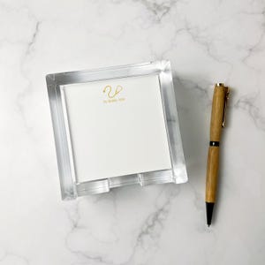 May include: A clear acrylic notepad holder with a white notepad featuring a gold stethoscope design and the text "Dr. Riddle, M.D." A wooden pen with gold accents rests beside the notepad holder.