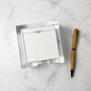 May include: Clear acrylic notepad holder with a white notepad featuring the initials "BWA". A wooden pen with gold accents and a black tip is positioned next to the notepad holder. The items are on a white marble surface.