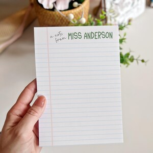 Puede incluir: Un bloc de notas blanco con l&iacute;neas horizontales azules y un margen vertical rosa. El texto "a note from MISS ANDERSON" est&aacute; impreso en verde y negro. El bloc de notas est&aacute; sostenido por una persona.