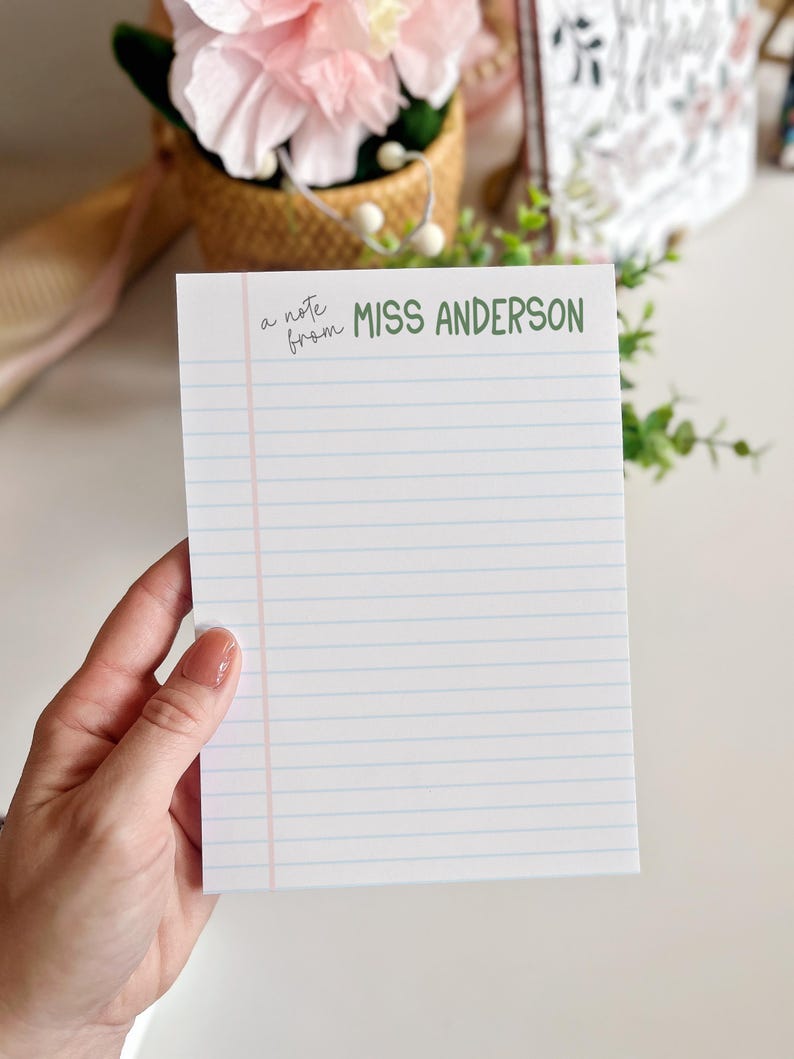 Puede incluir: Un bloc de notas blanco con l&iacute;neas horizontales azules y un margen vertical rosa. El texto "a note from MISS ANDERSON" est&aacute; impreso en verde y negro. El bloc de notas est&aacute; sostenido por una persona.