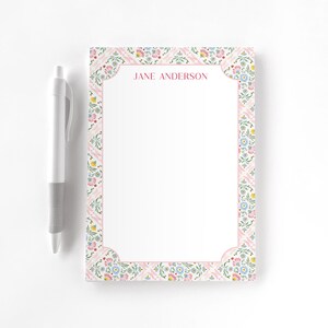 May include: A white notepad with a floral border and the text "JANE ANDERSON" in pink. A white and gray pen is positioned to the left of the notepad. The notepad has a pink border and a floral pattern.