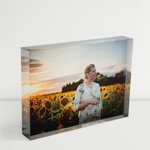 May include: A pregnant woman in a floral dress stands in a field of sunflowers at sunset. The sunflowers are in full bloom and the sky is a vibrant orange and pink.
