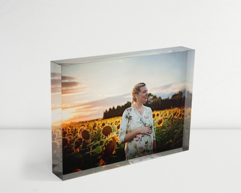 May include: A pregnant woman in a floral dress stands in a field of sunflowers at sunset. The sunflowers are in full bloom and the sky is a vibrant orange and pink.