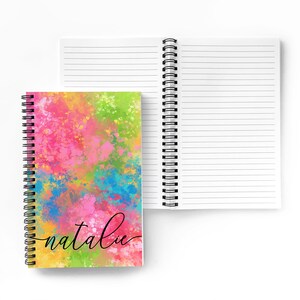 Puede incluir: Un cuaderno de espiral con un dise&ntilde;o abstracto colorido en la portada. El dise&ntilde;o presenta una mezcla de colores rosa, azul, amarillo y verde. El nombre "natalie" est&aacute; escrito en cursiva negra en la portada.
