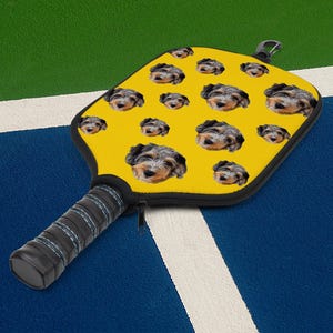 May include: Yellow pickleball paddle featuring a pattern of dog faces. The paddle has a black handle with a zipper and a black carabiner clip. The handle is wrapped in black grip tape with blue stitching. The paddle is designed for playing pickleball.