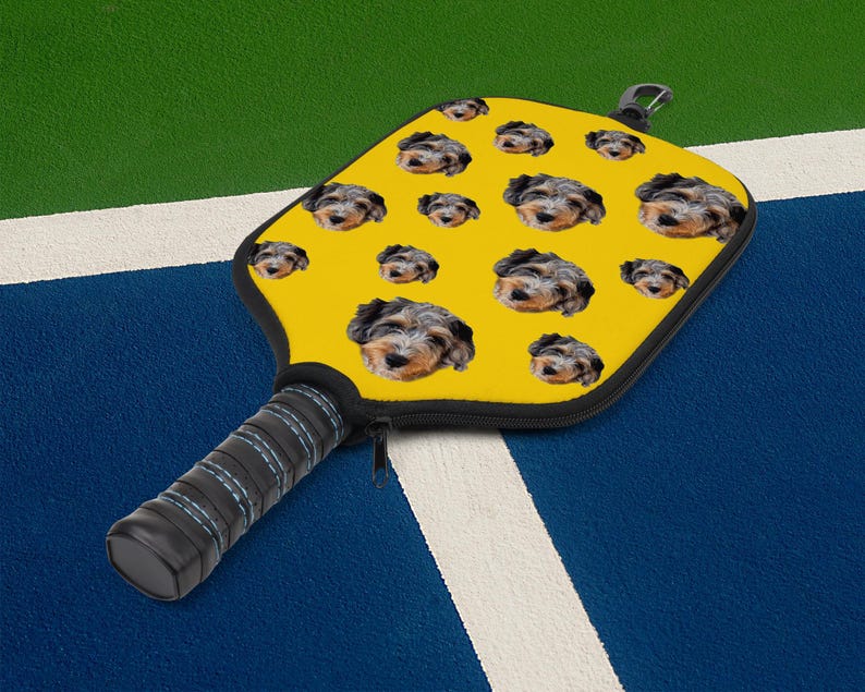 May include: Yellow pickleball paddle featuring a pattern of dog faces. The paddle has a black handle with a zipper and a black carabiner clip. The handle is wrapped in black grip tape with blue stitching. The paddle is designed for playing pickleball.