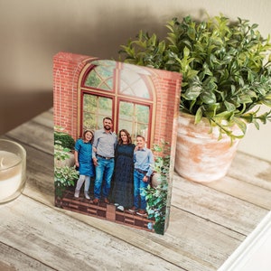 May include: A family of four poses in front of a brick building with a large arched window. The family is standing in a garden setting with green plants and flowers. The photo is printed on a clear acrylic block.