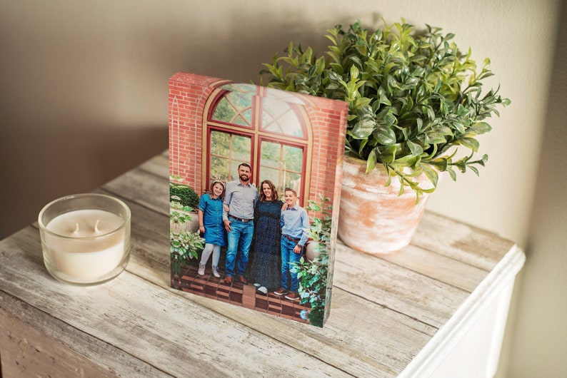 May include: A family of four poses in front of a brick building with a large arched window. The family is standing in a garden setting with green plants and flowers. The photo is printed on a clear acrylic block.